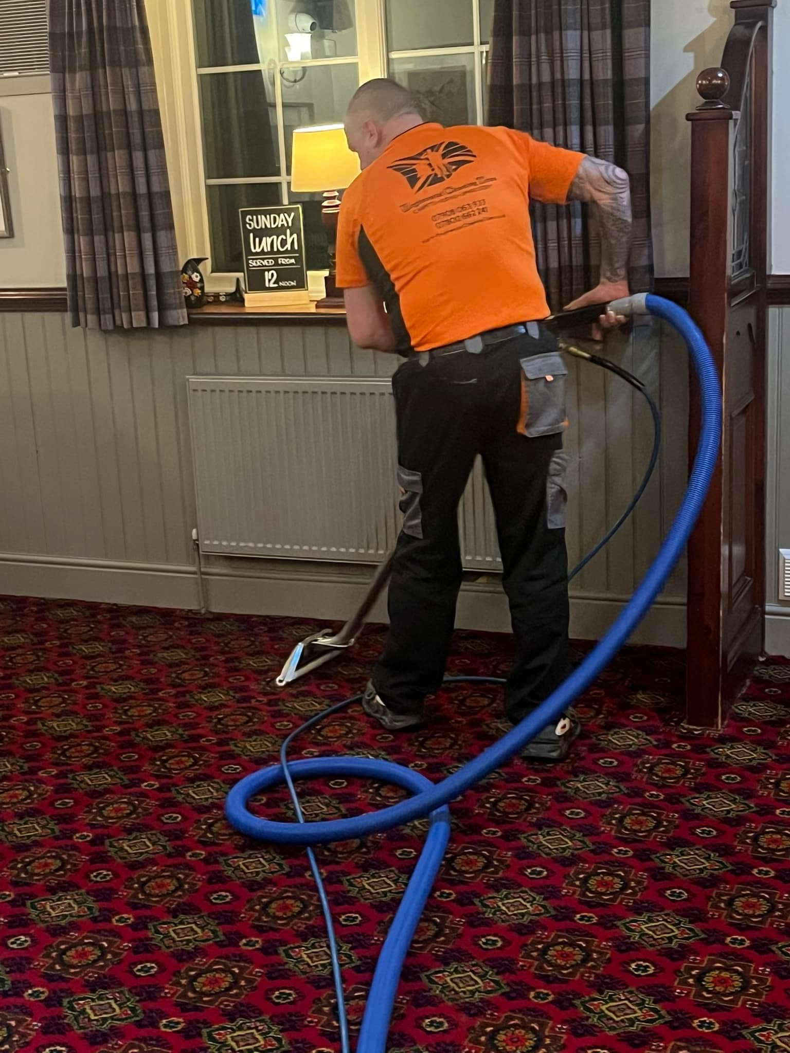 Pete in his orange Regimental Cleaning Team shirt cleaning a patterned commercial carpet with an extraction hose
